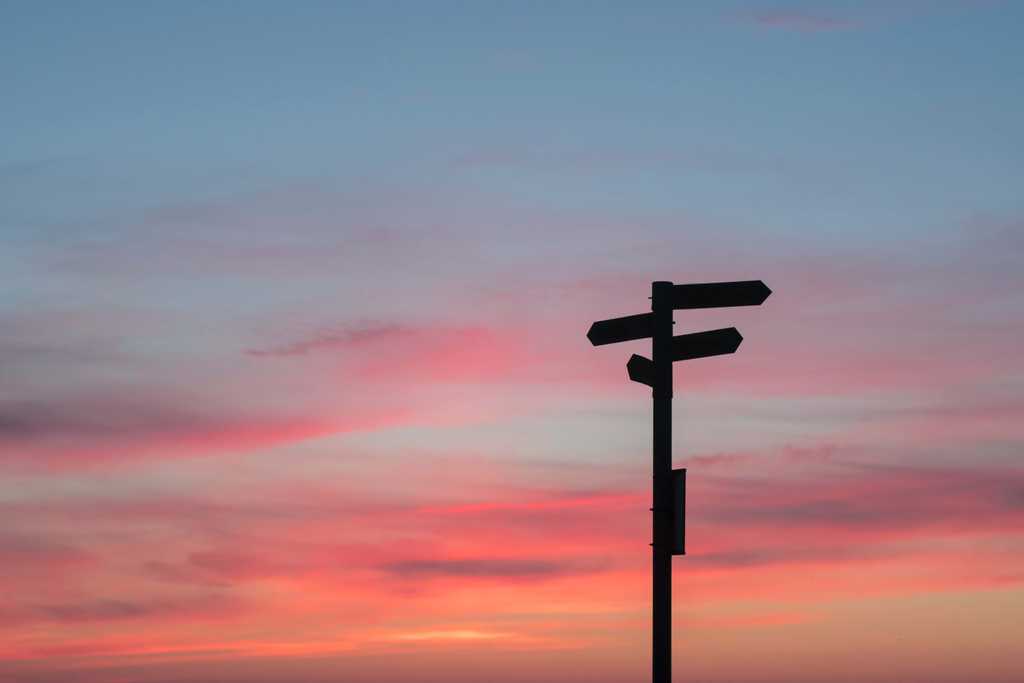 wooden sign points in multiple directions in front of pink sky during sunset