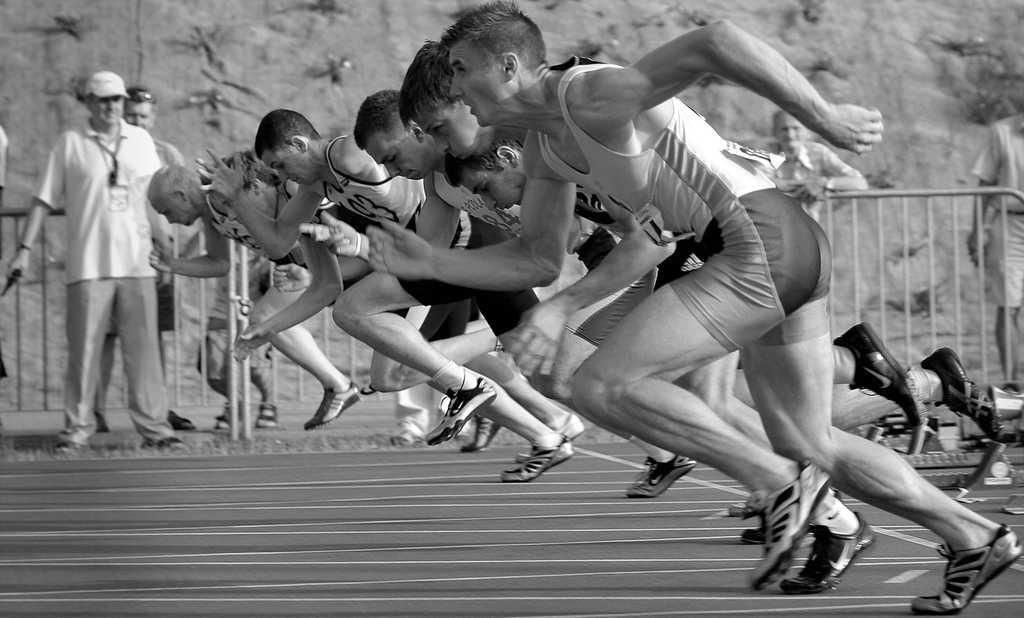 5 athletes in line starting a sprint at the starting line with two judges watching 