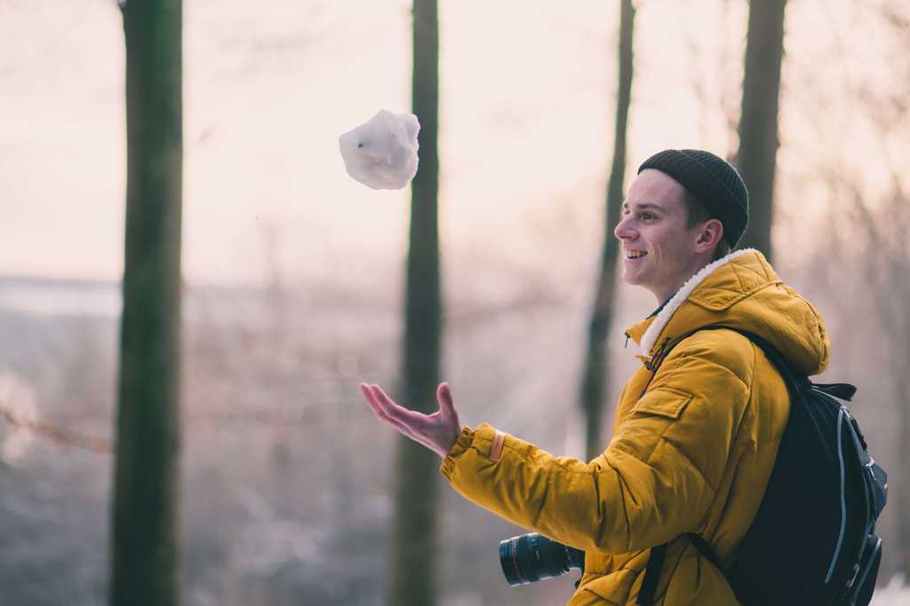 young man wearing a yellow jacket throws a snowball upward in the air in front of a snowy lightly forested environment